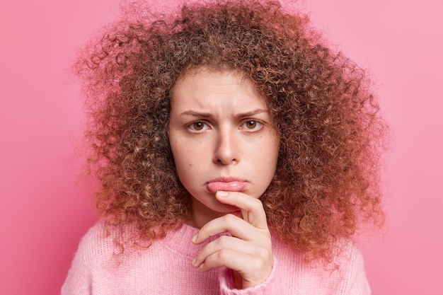 Free photo close up shot of displeased curly haired woman purses lips feels sad frowns face has discontent expression wears casual jumper isolated over pink wall. negative face expressions concept