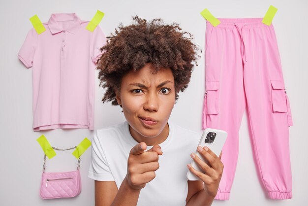 Close up shot of displeased woman looks angrily and points at you holds smartphone blames someone dressed in casual t shirt poses against white background with pink items of clothes. Hey listen to me