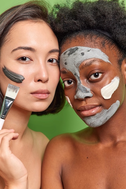 Free photo close up shot of diverse young women apply clay masks on face hold cosmetic brush look directly at camera stand shirtless indoor take care of complexion and skin isolated over green wall