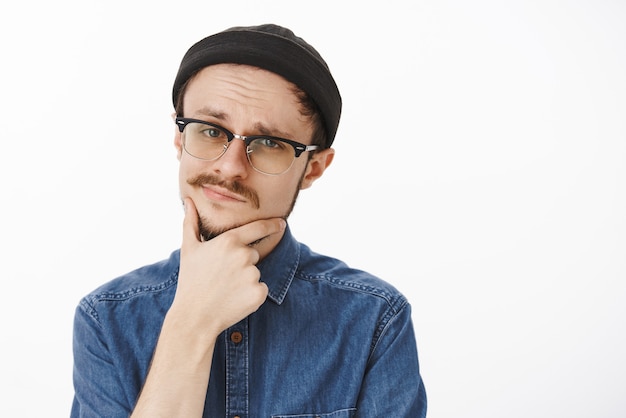 Close-up shot of doubtful smart suspicious handsome young guy with beard and moustache in black beanie and glasses touching chin and smirking squinting feeling uncertain and unimpressed