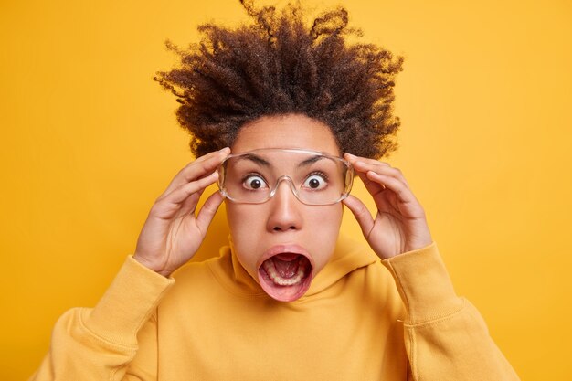 Close up shot of emotional super shocked ethnic woman popping eyes, keeps mouth wide opened notices terrible scene wears sweatshirt has hair standing up 