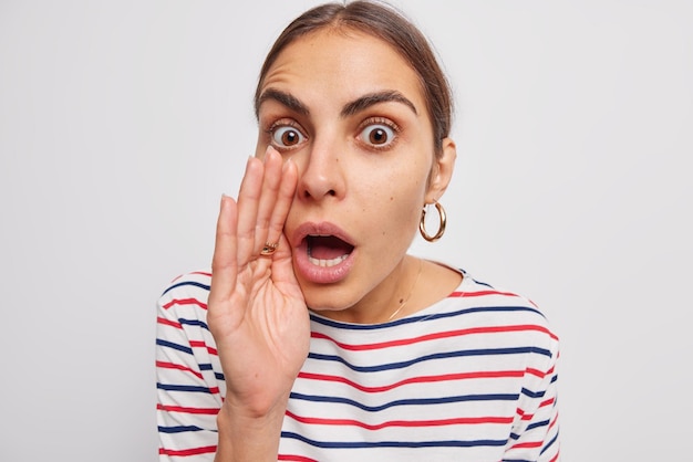 Close up shot of emotive shocked brunette young woman keeps palm near facetells secret spreads rumors stunned by amazing relevation dressed casually isolated over white background. Human reaction