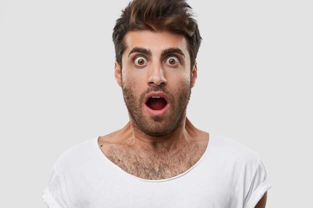 Close up shot of emotive surprised unshaven man with bugged eyes, wide opened mouth, has dark bristle, wears casual white t shirt