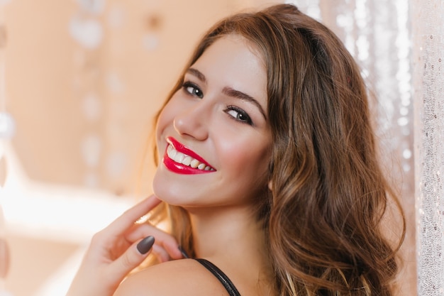 Close-up shot of European girl with dark curly hair and evening make-up smiling against silver wall.