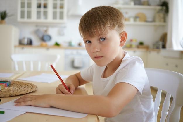 Close up shot of European schoolboy in white t-shirt sitting at wooden table drawing picture or doing homework with kitchen interior, looking , having serious facial expression