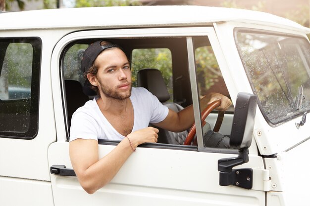 Close up shot of fashionable handsome young bearded model posing inside white jeep on driver's seat holding hand on steering wheel and looking with confident expression on his face