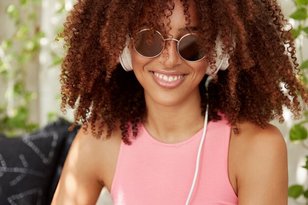 Free photo close up shot of female with bushy afro hairstyle, dark healthy skin, spends free time on listening music in headphones, enjoys favourite playlist, smiles happily. woman meloman poses indoor