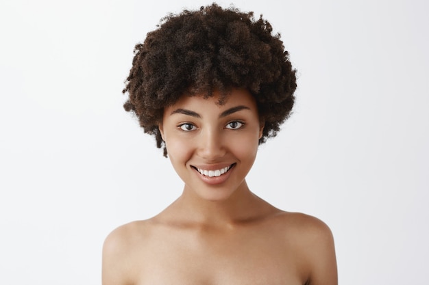 Снимок крупным планом женственной милой и натуральной афроамериканки с вьющимися волосами, стоящей обнаженной и широко улыбающейся, любящей и заботящейся о собственном теле