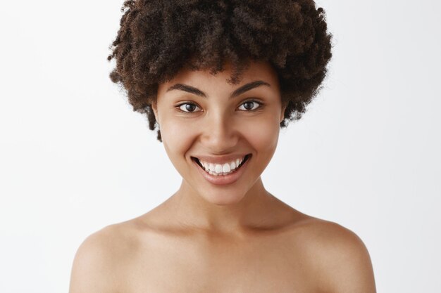 Крупным планом кокетливая и женственная уверенная в себе афроамериканка с широкой яркой улыбкой позирует обнаженной и довольна прекрасным состоянием кожи после косметологической процедуры