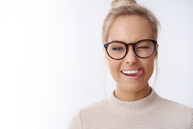 Close-up shot of flirty and playful caucasian blond woman in glasses combed haircut winking happily with positive attitude showing tongue enjoying weekends over white background