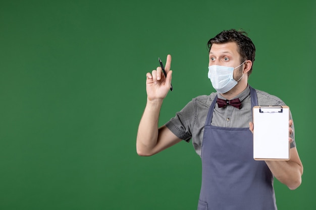 Close up shot of focused male waiter in uniform with medical mask and holding order book pen pointing up on green background