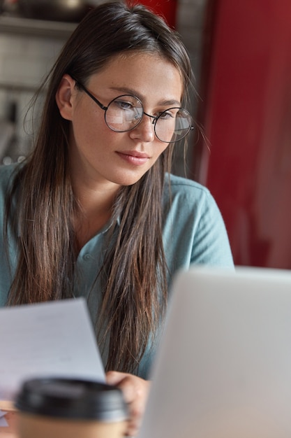 Close up shot of freckled woman in big round spectacles examines paper work, checks business project on laptop computer, drinks aromatic coffee, poses indoor, reads recommendation from client