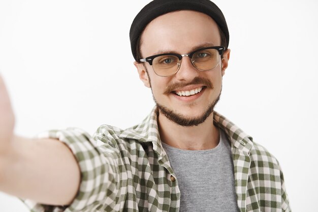 Close-up shot of friendly handsome neat guy in black trendy beanie and glasses with moustache smiling broadly while taking selfie