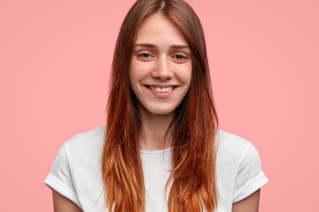 Close up shot of friendly looking female with positive expression, has charming smile, stands against pink background. people and emotions concept