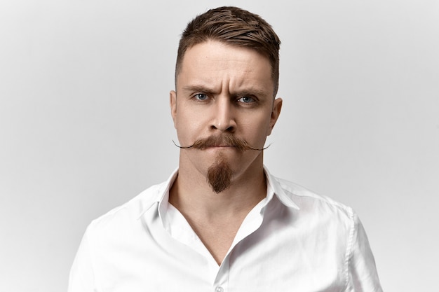 Close up shot of frustrated young man with stylish haircut, mustache and stubble frowning his eyebrows and pursing lips, having uneasy perplexed expression, being worried about problems at work