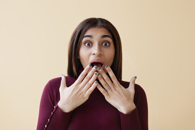 Close up shot of funny amazed young African American female wearing bright nail polisher expressing surprised emotions, covering wide opened mouth with hands. Astonishment, shock and excitement