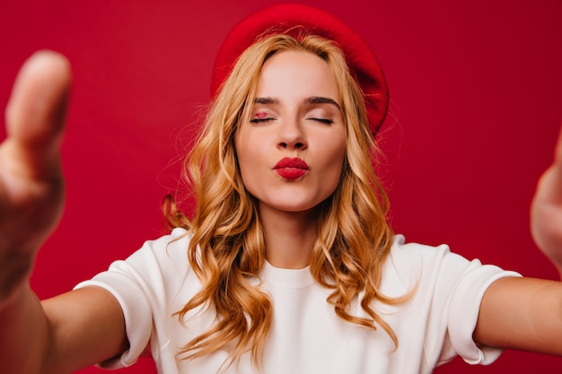 Close-up shot of funny french girl posing with kissing face expression on red wall. cute blonde woman wears trendy beret.