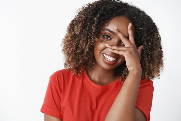 Close-up shot of funny tender and feminine dark-skinned woman with curly haircut close half of face with hand and smiling, giggling enjoying great amusing company over white background. Copy space