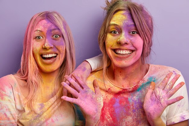 Close up shot of glad smiling women being in high spirit, smile broadly, show palms, have dirty faces and palms with Holi colors, have fun indoor, isolated over purple wall. Celebration