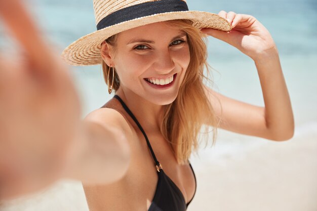 Close up shot of good looking female tourist enjoys free time outdoor near ocean on beach,  during leisure on sunny summer day, poses for selfie.
