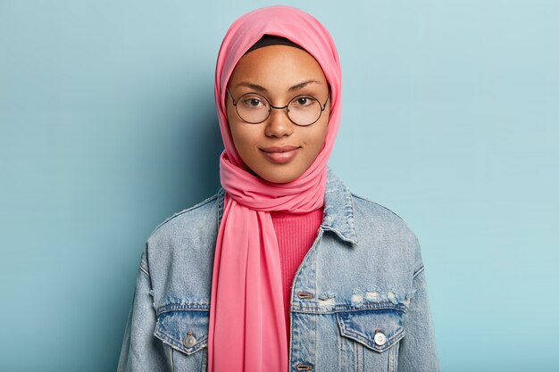 건강한 피부를 가진 좋은 찾고 여자의 총을 닫고, 투명 안경, 머리에 분홍색 스카프, 파란색 벽 위에 절연 진 재킷을 착용하고 자신감이 있습니다. 종교 개념