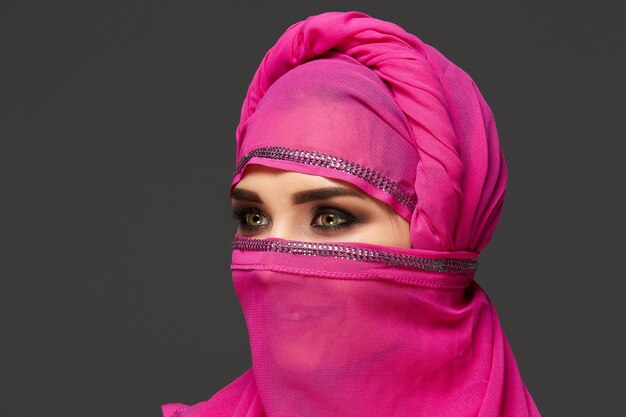 Close-up shot of a good-looking young female with an expressive smoky eyes wearing the chic pink hijab decorated with sequins. She has turned her head and looking away on a dark background. Human emot