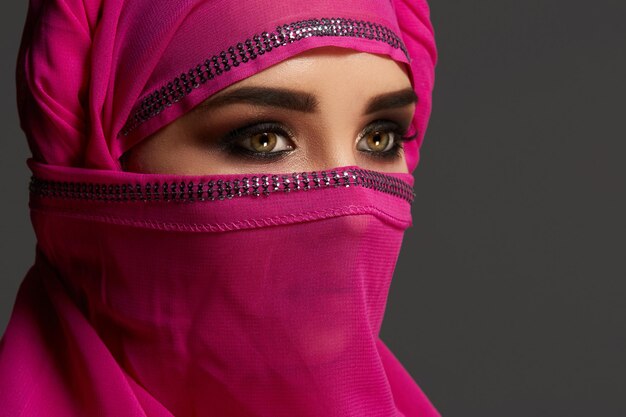 Close-up shot of a good-looking young girl with an expressive smoky eyes wearing the chic pink hijab decorated with sequins. She is posing at the studio and looking away on a dark background. Human em