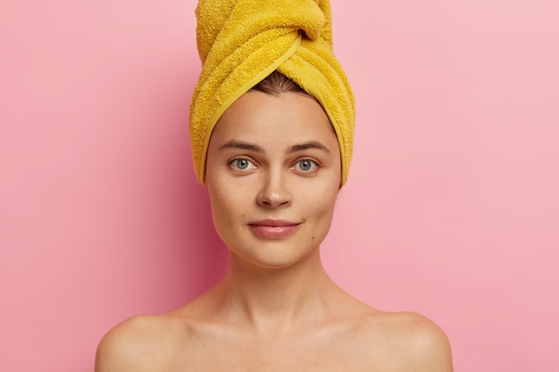 Close up shot of gorgeous fresh European woman with towel on head, has clean face, healthy skin, stands shirtless, takes shower, going to apply makeup, has natural beauty. Body care concept.