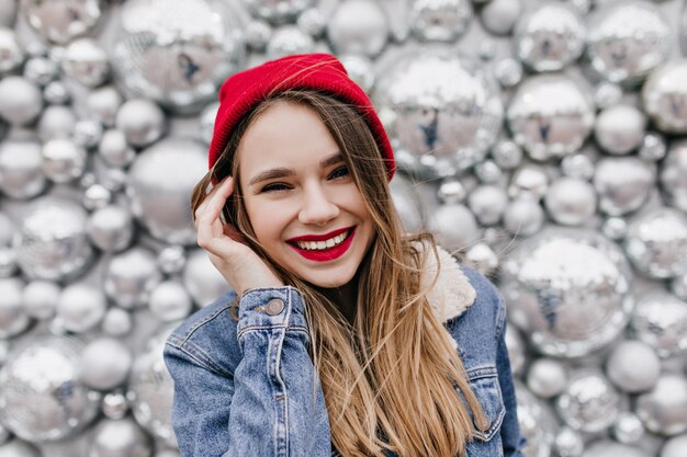 Close-up shot of gorgeous white woman with long hair smiling on sparkle wall in cold day. Ecstatic girl in red hat and denim jacket laughing during photoshoot.