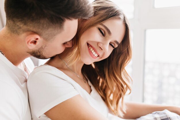 Close-up shot of gorgeous young lady enjoying leisure time with beloved man. Indoor portrait of laughing curly girl chilling with husband at home.