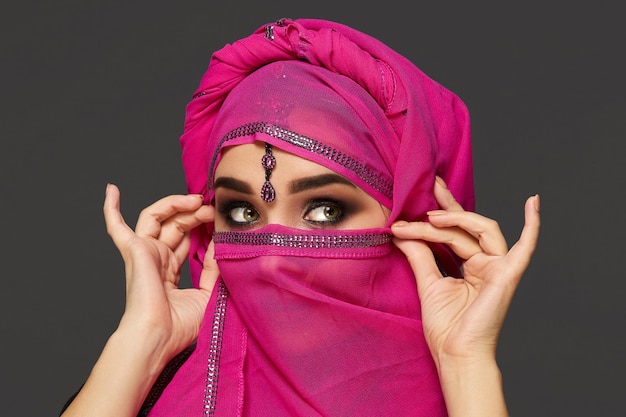 Close-up shot of a gorgeous young woman with an expressive smoky eyes wearing the chic pink hijab decorated with sequins and jewelry. She is holding the shawl with her hands and looking away on a dark