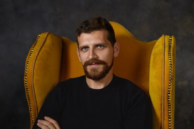 Close up shot of handsome unshaven male with blue eyes, fuzzy beard and mustache relaxing indoors, sitting isolated in stylish armchair. People, style, interior design and furniture concept