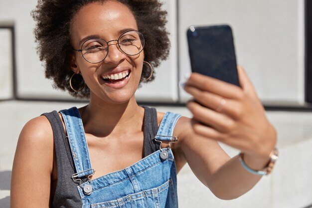 Close up shot of happy African American woman holds cellular in front, smiles broadly, takes selfie portrait, being in high spirit, enjoys outdoor rest, dressed in fashionable summer clothes. Leisure