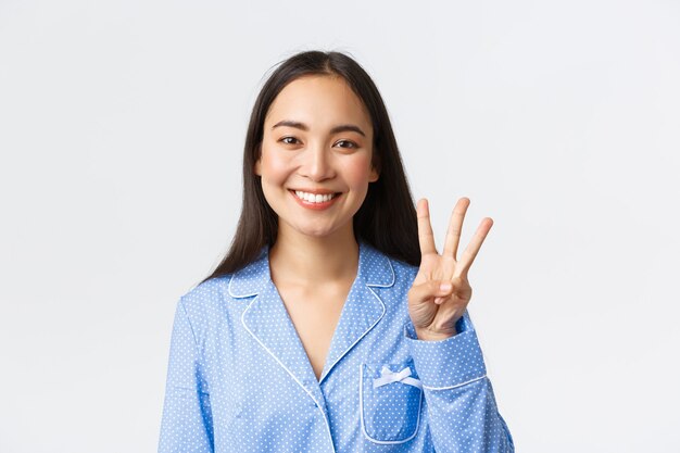 Close-up shot of happy attractive asian woman in blue pyjama showing three fingers and smiling white teeth, explain main rules or making order, standing white background delighted.