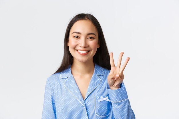 파란 잠옷을 입은 행복한 매력적인 아시아 여성의 클로즈업 샷은 세 손가락과 하얀 치아를 보여주고, 주요 규칙을 설명하거나 주문을 하고, 흰색 배경을 기뻐합니다.