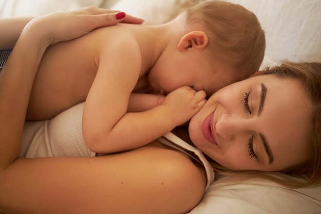 Закройте снимок счастливой очаровательной молодой матери, лежа на кровати со своим сонным маленьким ребенком на груди. Красивая мама и младенец обнимаются в спальне. Концепция любви, счастья, материнства и воспитания