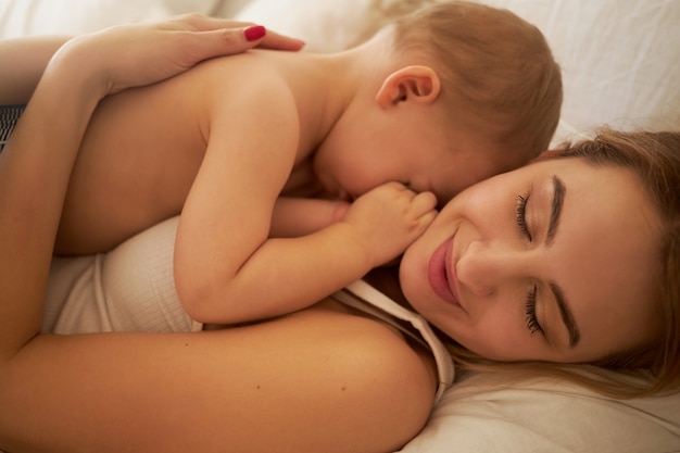 彼女の眠そうな小さな子供を胸にベッドに横たわっている幸せな魅力的な若い母親のショットをクローズアップ。寝室で抱きしめるかわいいお母さんと幼児。愛、幸福、出産、子育ての概念