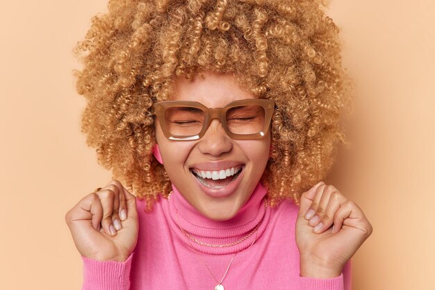 幸せな縮れ毛の女性のクローズアップショットは手を振る笑顔は広く目を閉じたままでエネルギッシュな眼鏡をかけていると感じ、ベージュの背景の上に分離されたカジュアルなピンクのタートルネックは素晴らしいニュースを聞く