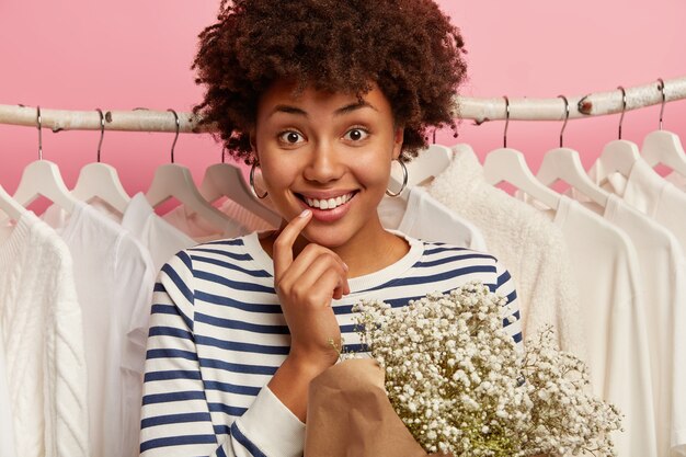 Close up shot of happy curly haired woman stands near white clothes on stores rails, dressed in sailor striped jumper, holds beautiful bouquet