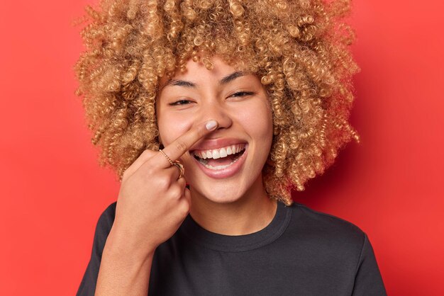 Close up shot of happy curly haired woman touches nose feels joyful smiles broadly tries to amuse someone dressed in casual black t shirt isolated over vivid red background has playful expression