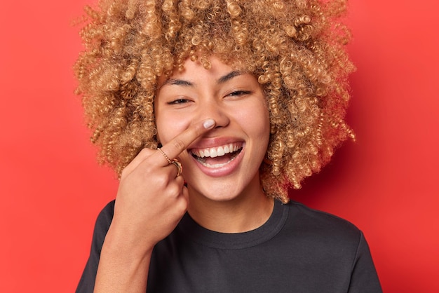 Close up shot of happy curly haired woman touches nose feels joyful smiles broadly tries to amuse someone dressed in casual black t shirt isolated over vivid red background has playful expression