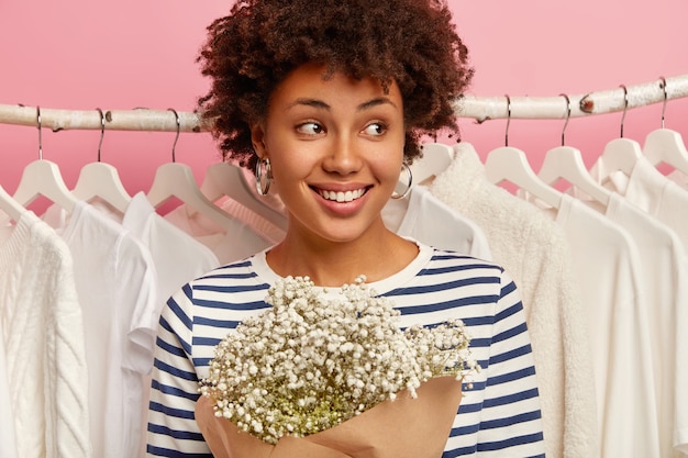 Close up shot of happy curly woman dressed in striped outfit, stands near clothes racks, looks aside with smile. Shopping and spring cleaning