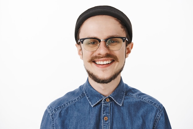 Close-up shot of happy delighted charming young bearded man with moustache in glasses and black trendy beanie smiling joyfully and laughing feeling satisfied and lucky