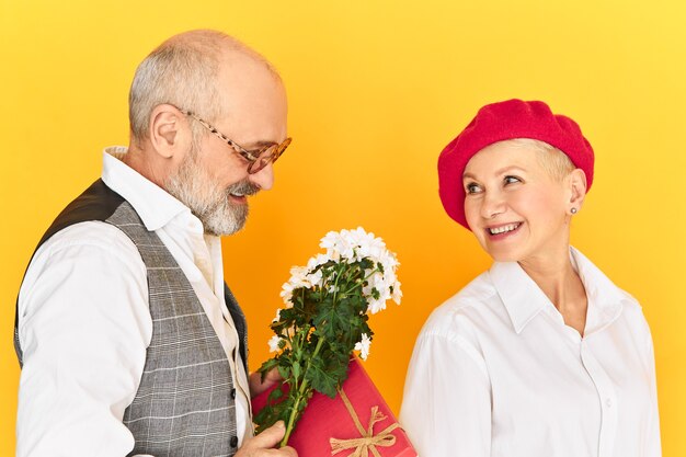 Close up shot of happy elderly couple in stlyish elegant clothes celebrating marriage anniversary, still in love with each other. Handsome male pensioner giving flowers to his charming woman