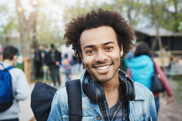 Close-up shot of happy emotive young african-american guy with afro hairstyle and bristle, smiling broadly while wearing denim coat and backpack, walking across park during festival