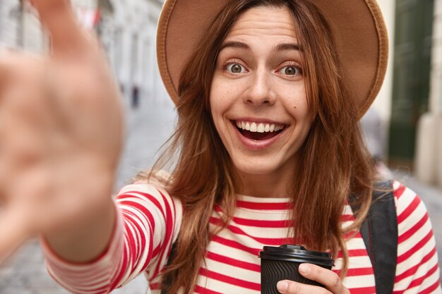 Close up shot of happy female traveler has hands outstretched at camera, makes selfie portrait