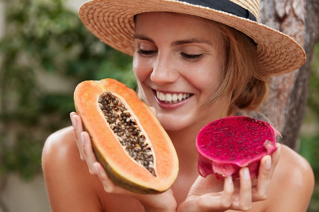 Close up shot of happy female with cheerful expression motivates you eat healthy food, holds papaya and dragon fruit.