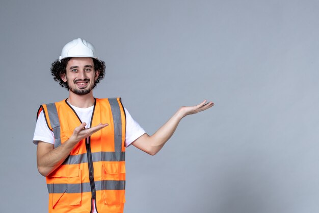 Close up shot of happy male constructor in warning vest wearing safety helmet and pointing something on the left side over gray wave wall