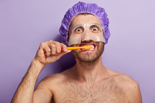 Крупным планом счастливый мужчина чистит зубы утром, наносит косметическую маску и носит шапочку для душа
