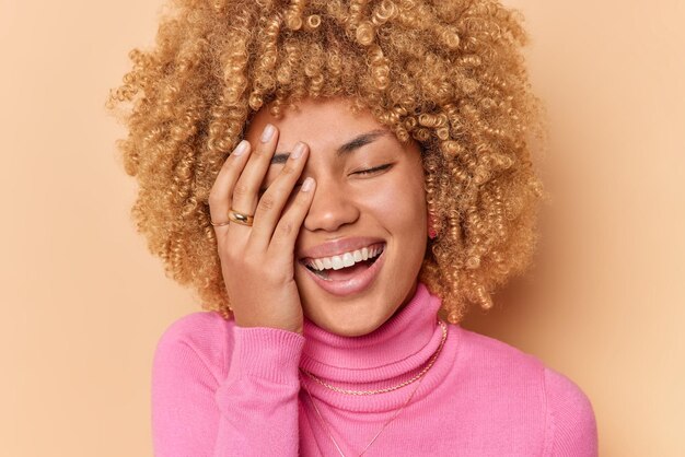 Close up shot of happy woman makes face palm smiles broadly shows white perfect teeth feels glad closes eyes from satisfaction wears pink turtleneck isolated on beige background has joyful expression